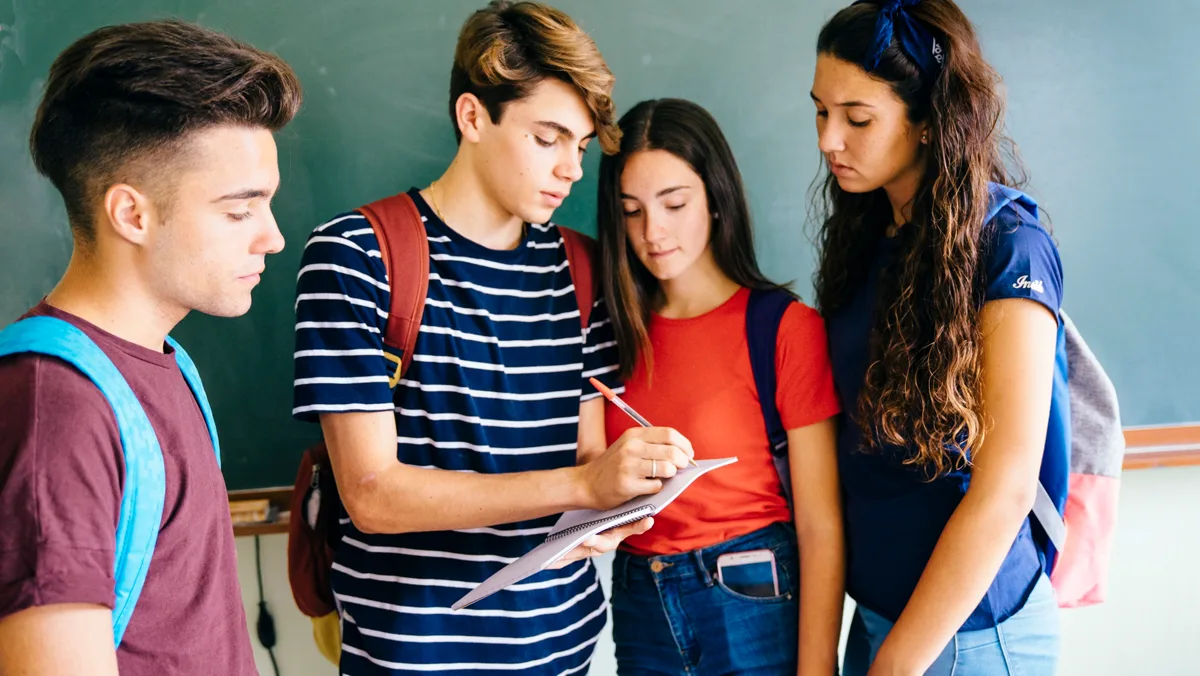 Four students in front of a chalkboard looking at a notebook one of them is holding