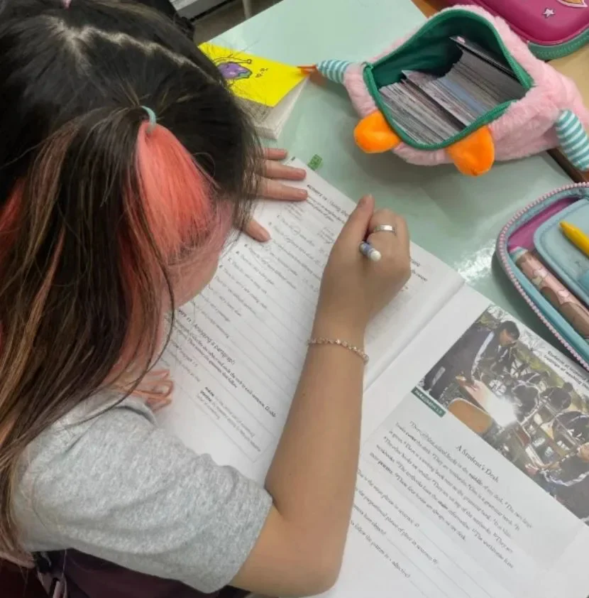 A student's hand writing sentences directly in an English textbook, with the example sentences open alongside