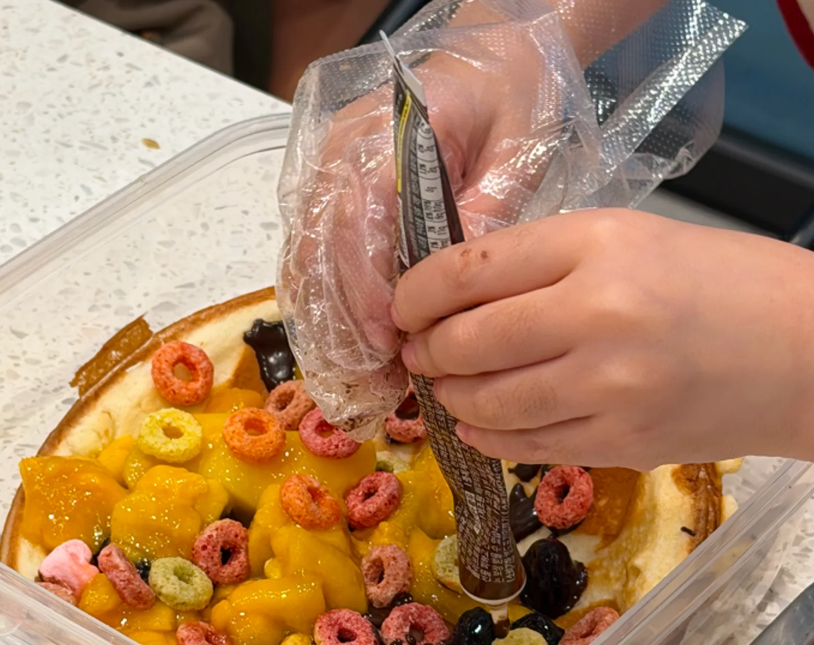 A gloved hand piping chocolate onto a tart topped with cereal and mango