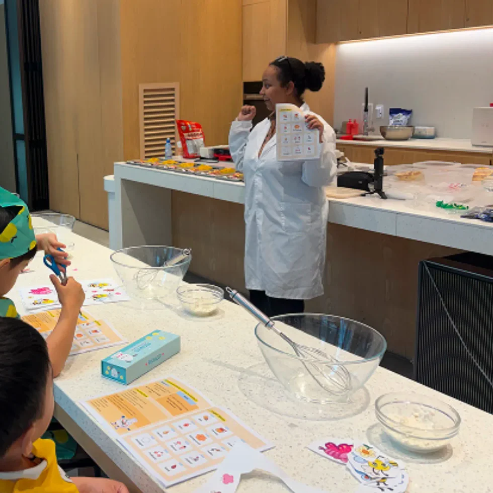 An instructor holds up a picture card while children look at worksheets on their desks in a Food & Science class