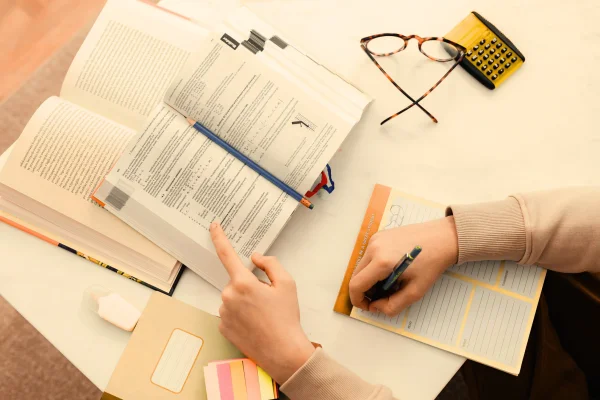 A student following the text in a book with a finger while writing notes in a notebook