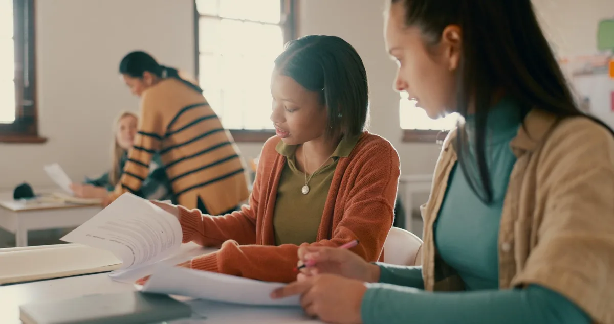 Two students at a classroom desk looking through papers together